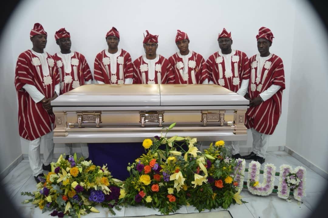 Pallbearers in red and white traditional attire standing behind a gold casket with flower tributes